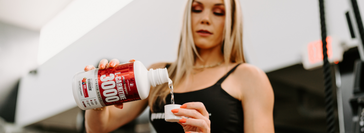 Woman pouring liquid L-carnitine supplement into a measuring cap in a gym setting, highlighting fitness and performance nutrition.