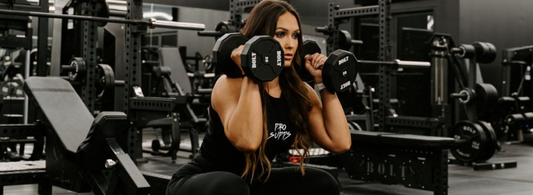 Woman performing dumbbell shoulder press in a gym surrounded by weight machines and strength training equipment.